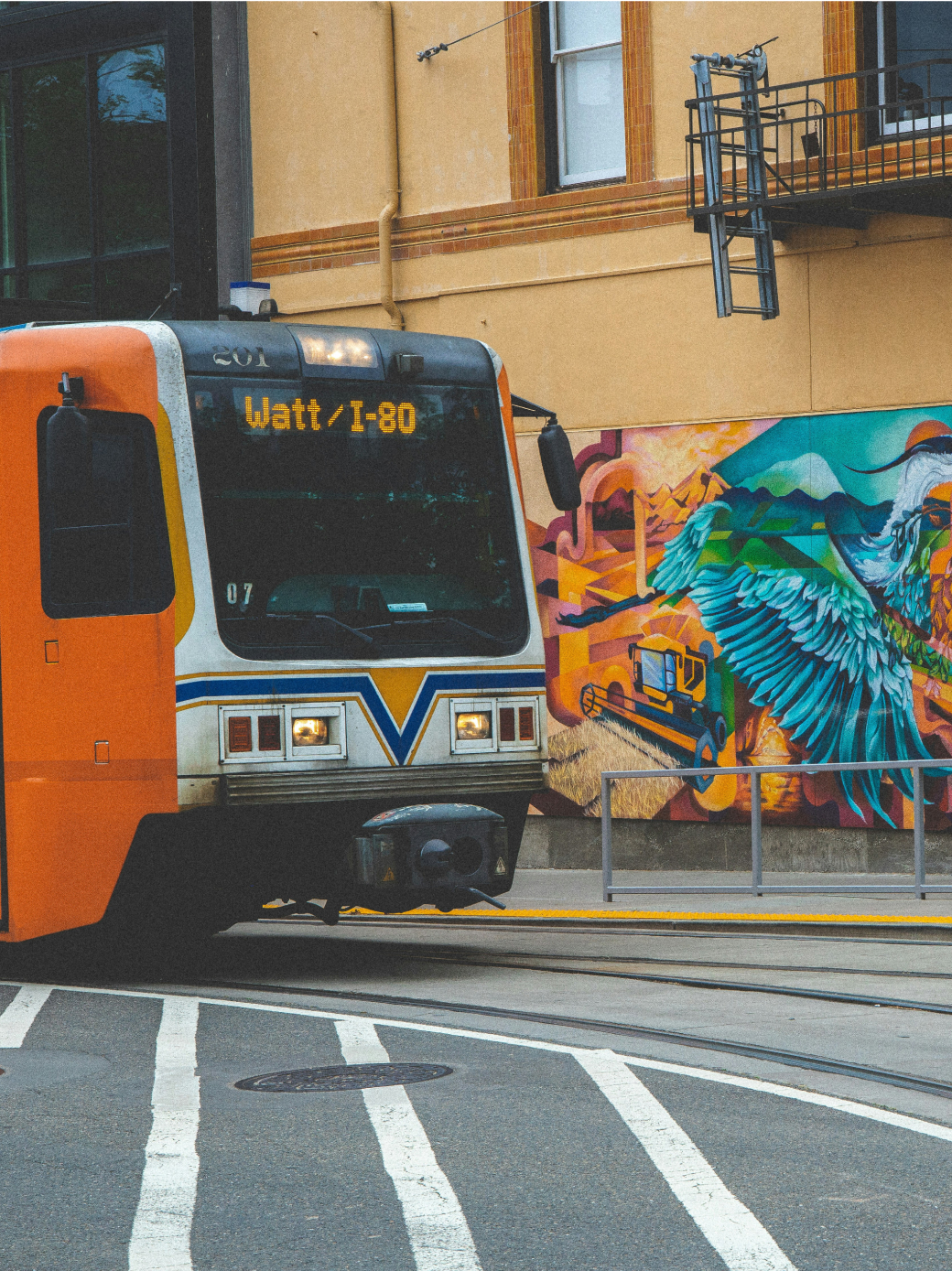 Orange light rail in Sacramento in front of a building with colorful mural.