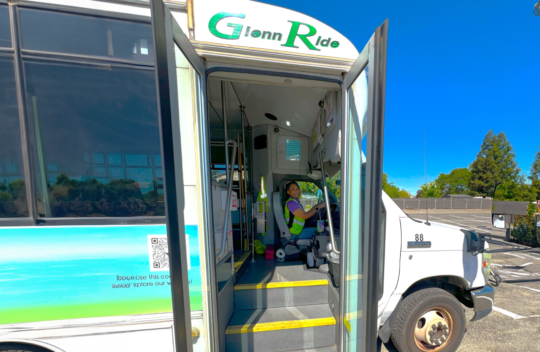 An accessible bus with doors open, showing a smiling bus driver inside. The bus is labeled “Glenn Ride” and parked in a sunlit lot with trees in the background.